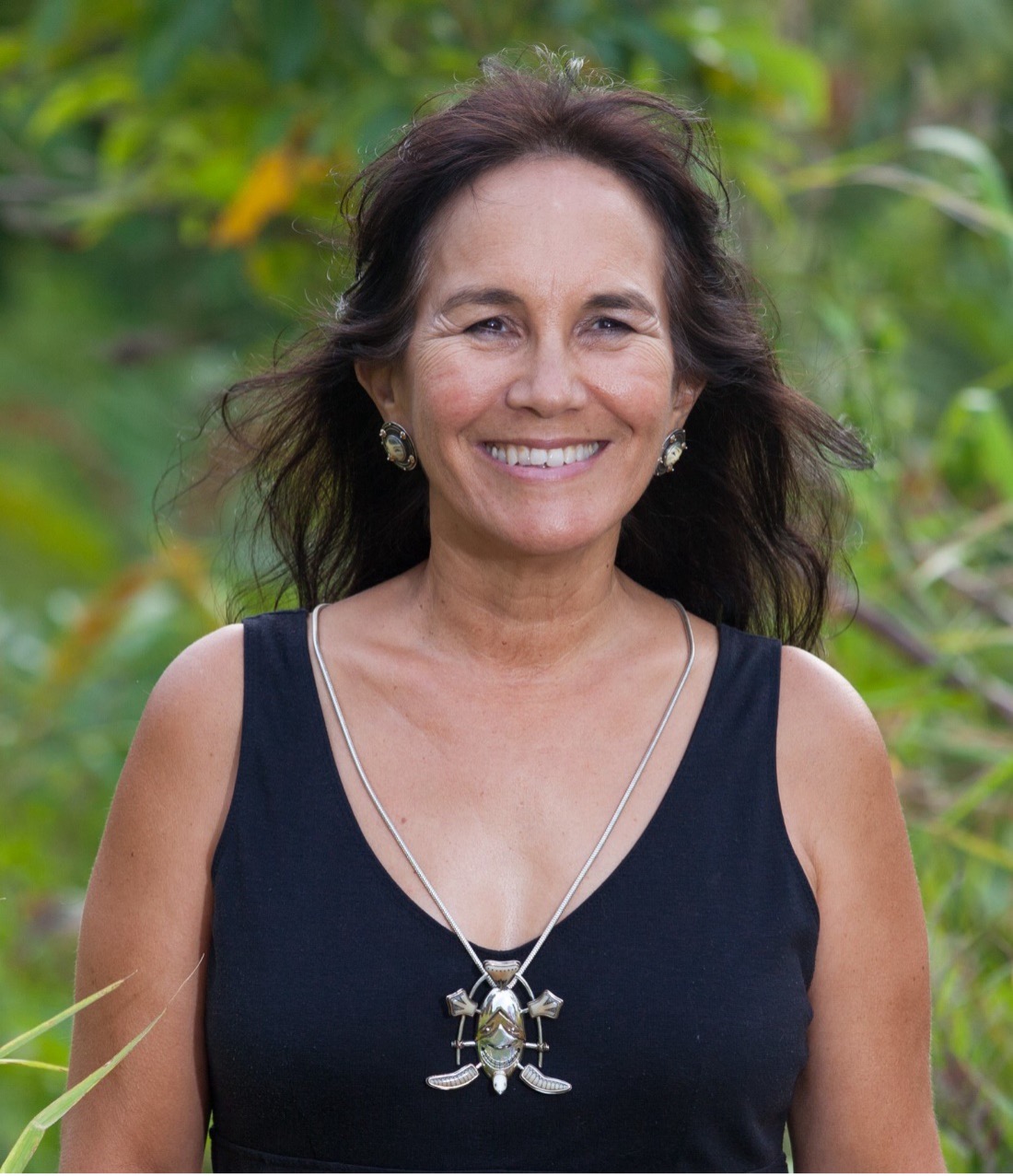 Photo of artist Denise Wallice wearing a black shirt with a silver necklace on standing outside smiling with green vegetation in the background
