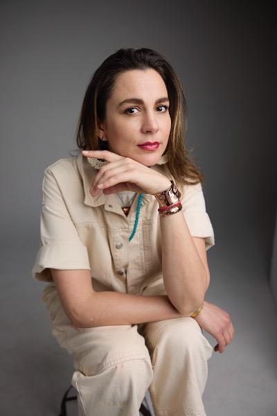 Image of artist Sáandlaanaay/Kimberly Fulton Orozco wearing a tan jumpsuit knelling down with left hand raised to their chin on a gray toned background
