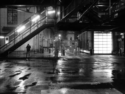 Black and White photograph. Image of a street in Chicago with an above ground entrance to the subway "L" station. The photograph is taken at night and the street lights are bright, reflecting off the wet street, lighting the cross walk lines on the pavement.