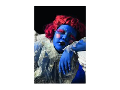 Color photograph. A close-up portrait of a feminine looking person leaning about a white column, their elbow resting and their head leaning against their arm. Their whole body is painted all blue and wearing a red wing. Their make-up is dramatic and draws attention. They are wearing long bright red eyelashes and red eyeshadow that matches their wing. They are also wearing a see-through white lace dress.