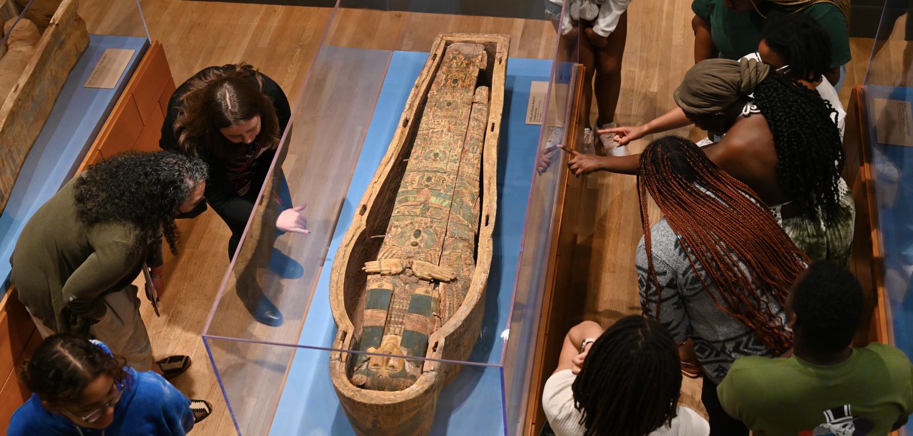 A group of adults gather around an ancient Egyptian coffin and point at images on the side