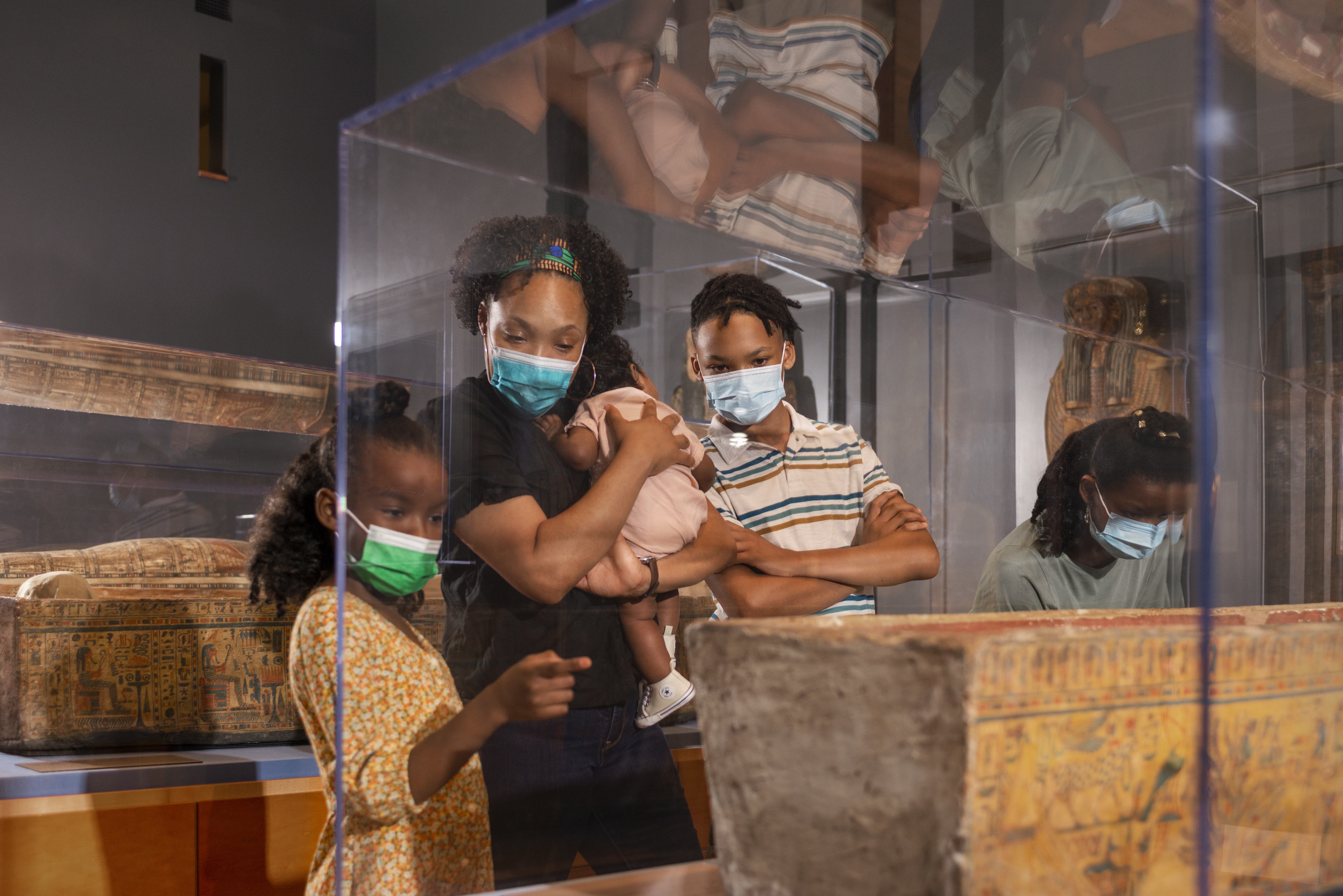 Photo of a homeschool family: a young girl, mother holding a baby, and young boy, all wearing face masks, observe an ancient Egyptian coffin