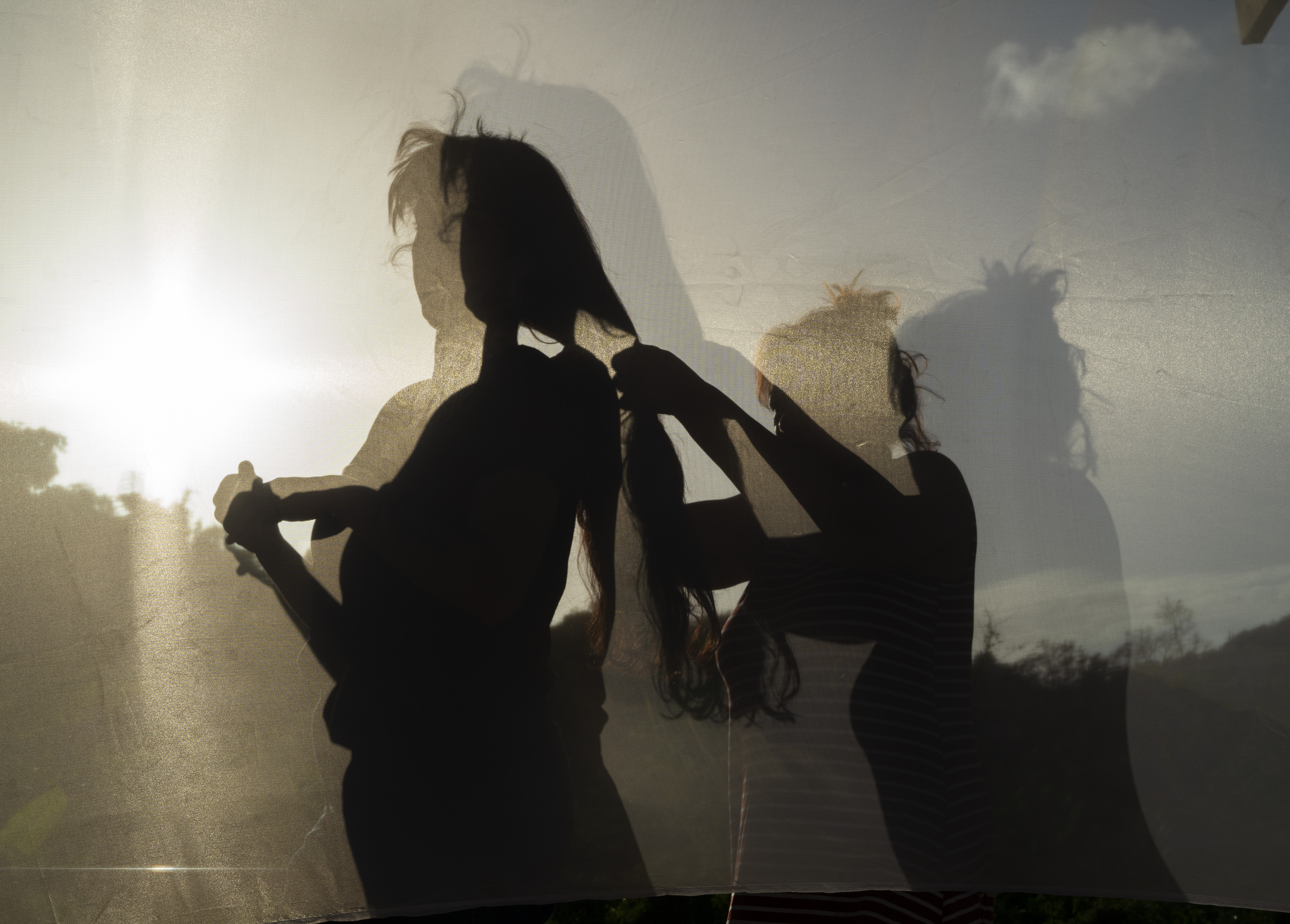 Color photo of two figures and their shadows. The left figure is standing while the figure behind them braids their hair.
