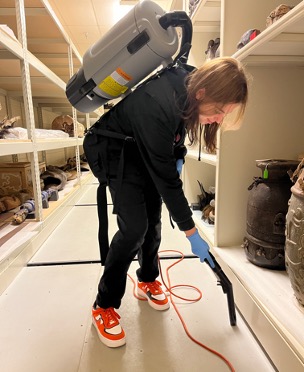 Audra Buffington is leaning down with a backpack vacuum on her back cleaning the museum storage area 