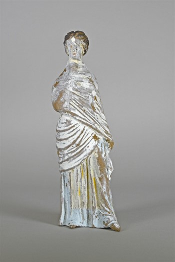 A painted Tanagra is shown on a gray background. The female form is heavily draped with pigments of white, yellow and blue on her dress 