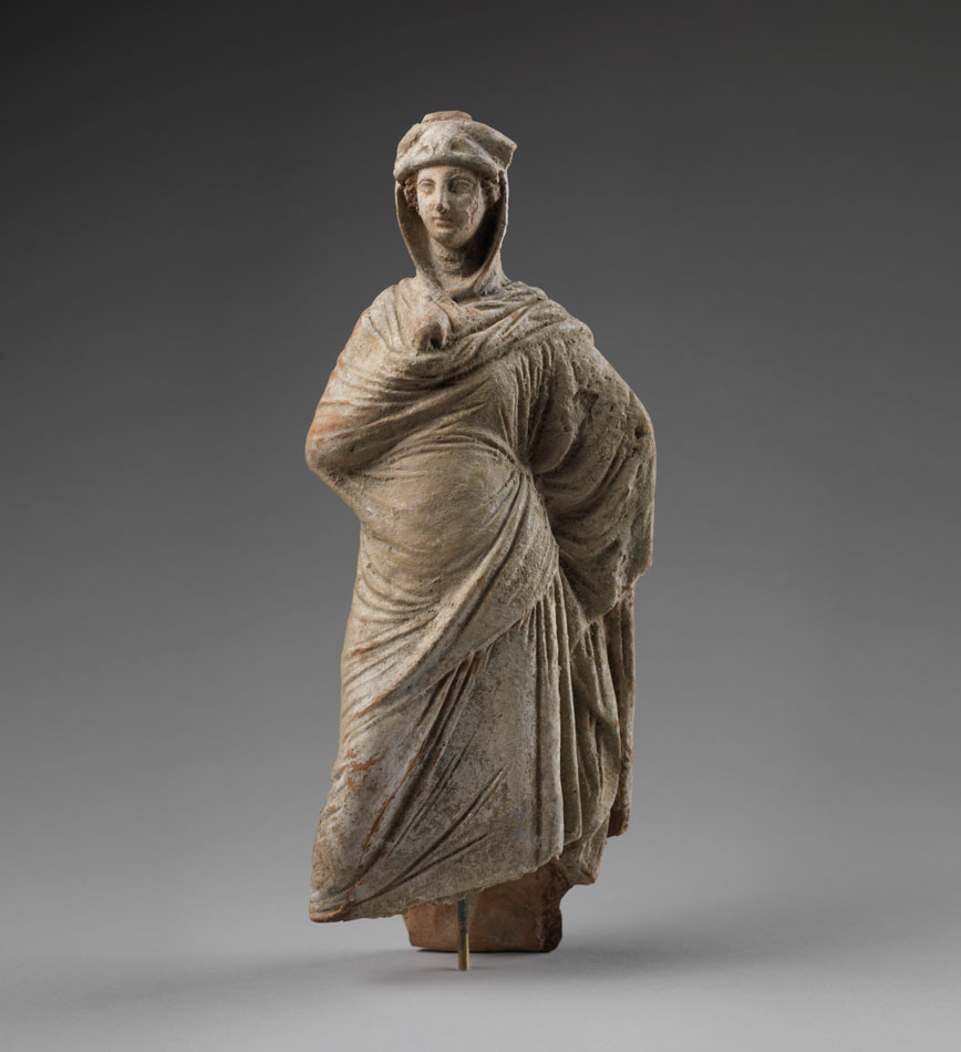 Small Tanagra figurine of a draped woman made in terracotta is shown on a faded background of gray and white. The figurine is missing it's base.