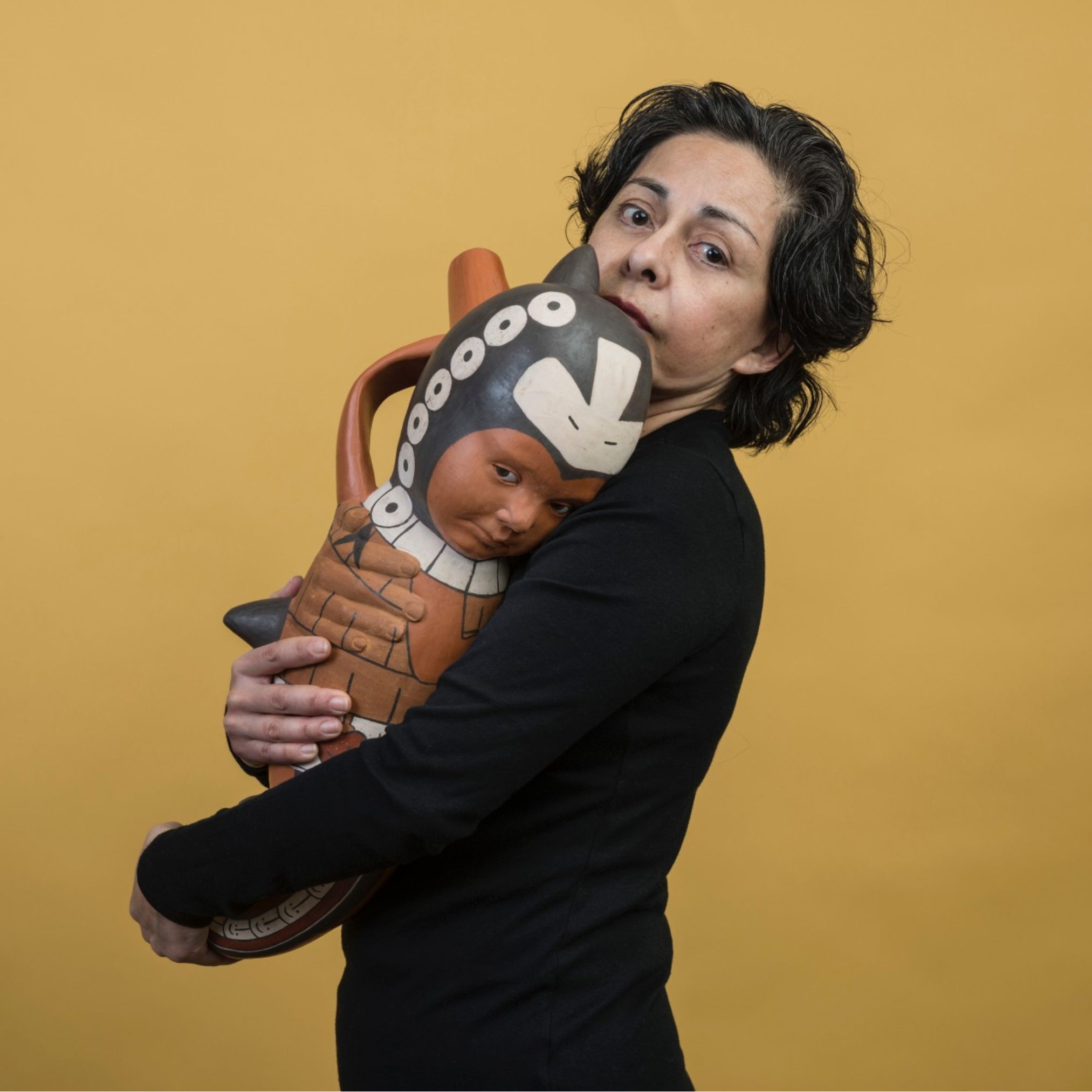 Kukuli Velarde photo on a yellow background holding one of her ceramic sculptures 