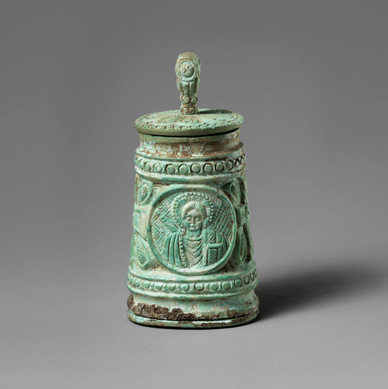 This object is a small vessel of carved bone, standing at only 3 1/8 inches tall. It is composed of a lid with a delicate, knob-shaped handle, a main body, which is hollow on the inside, and a disk-shaped base. The vessel’s lid and body are both intricately carved. The cone-shaped body of the vessel bears two portraits in roundel-shaped frames and two crosses with palmette-shaped blooms.