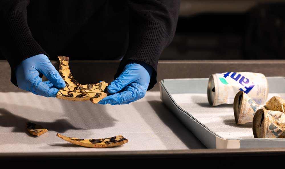 Hands with blue gloves hold together sherds next to a tray of Fanta cans