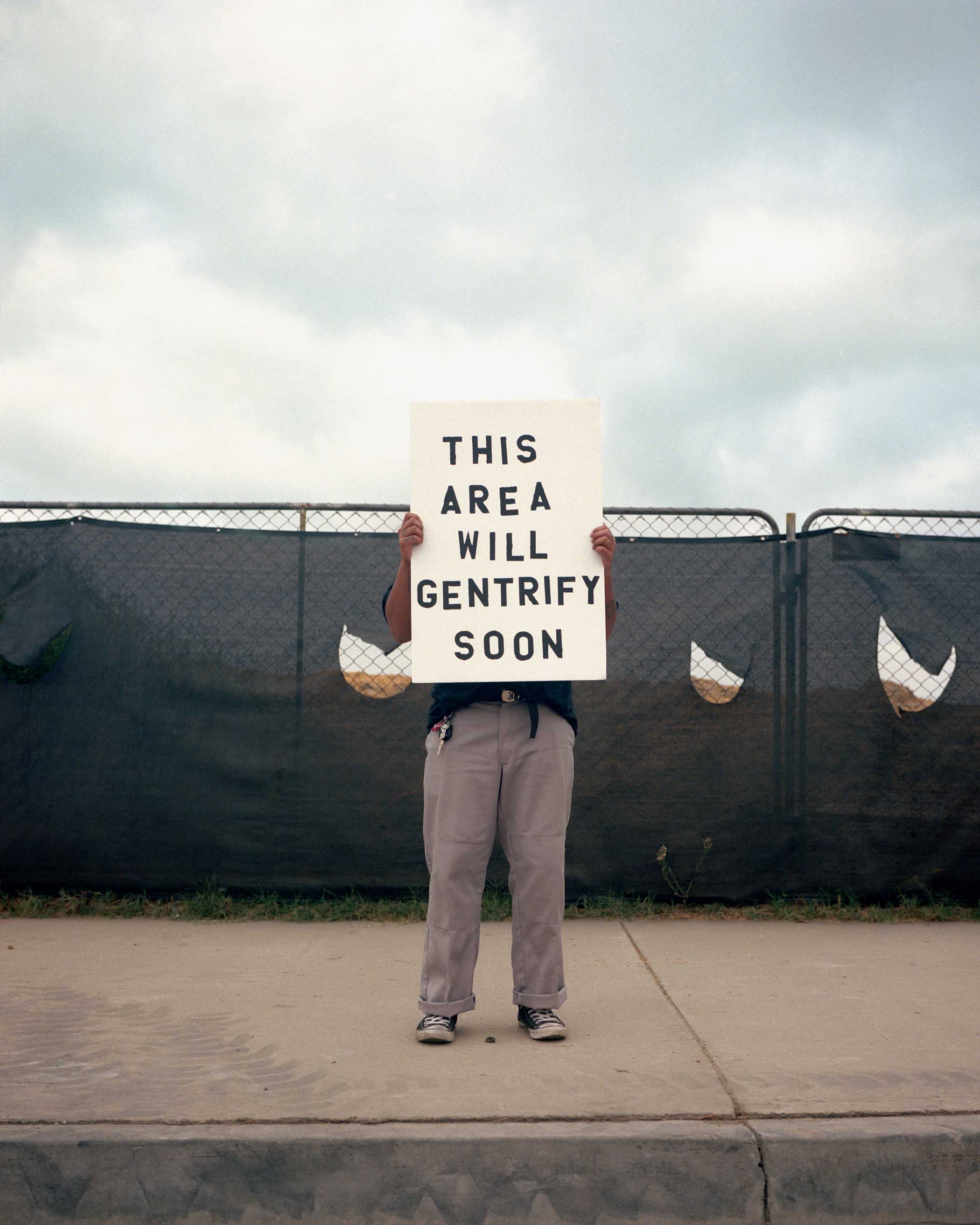 William Camargo, photography "We Gonna Have to Move Out Soon Fam!"  showing a person standing on a sidewalk infront of a fence. The white sign is covering the person's face and reads "This area will gentrify soon"