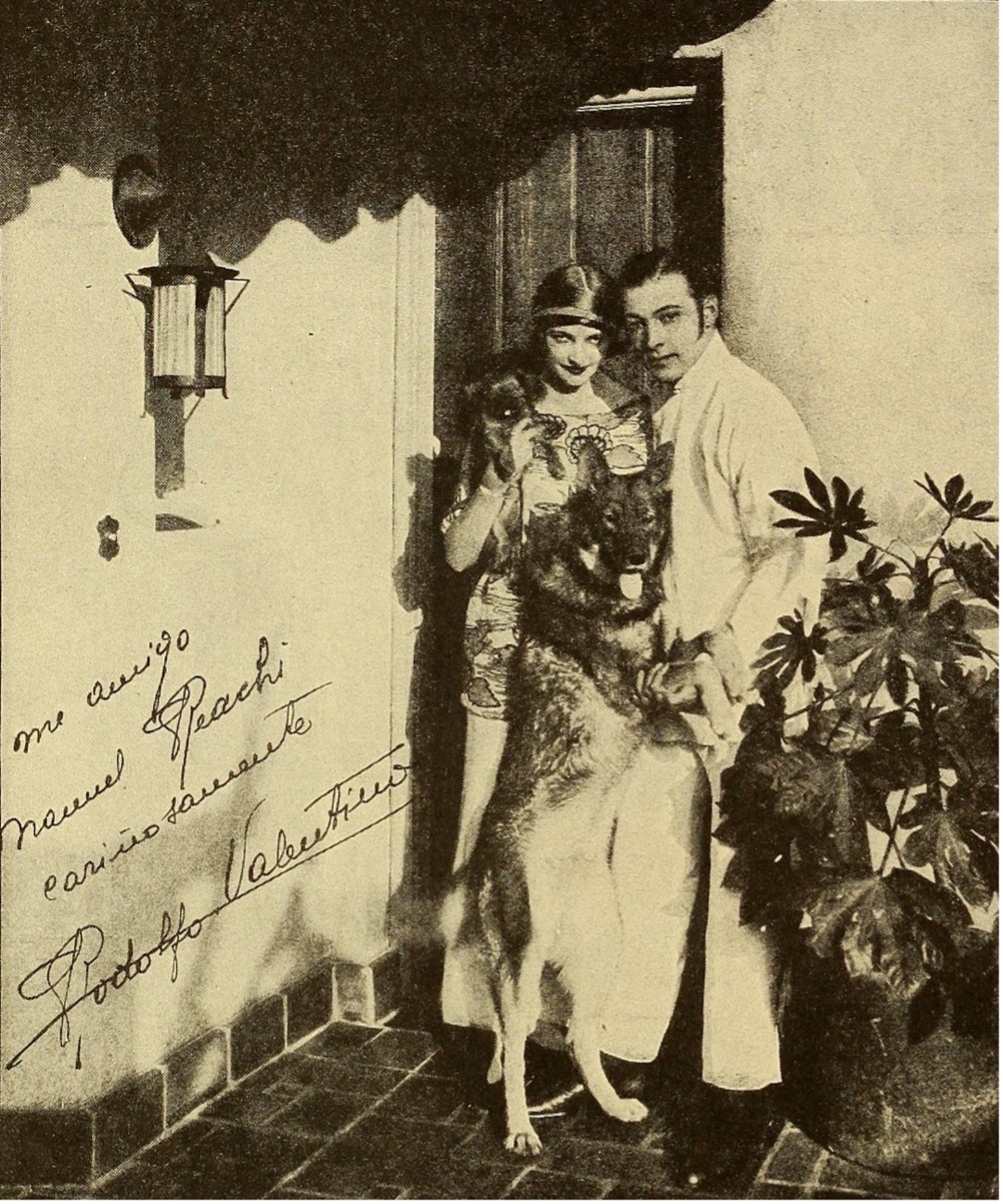 1930s photo of Natacha Rambova and Rudolph Valentino with their two dogs standing outside of a building, text is hand-written on the image