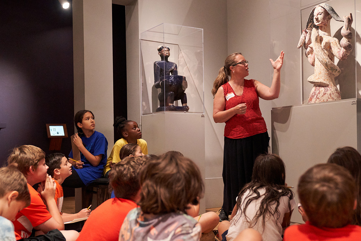 museum educator talking and gesturing to a sculpture of Mami Wata in front of a group of seated school-aged children.