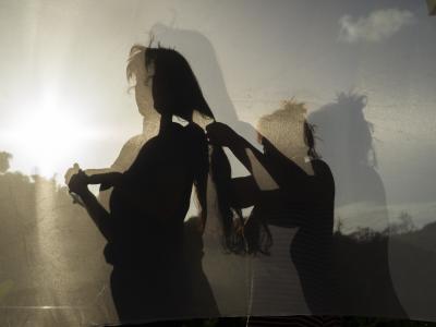 Color photo of two figures and their shadows. The left figure is standing while the figure behind them braids their hair.