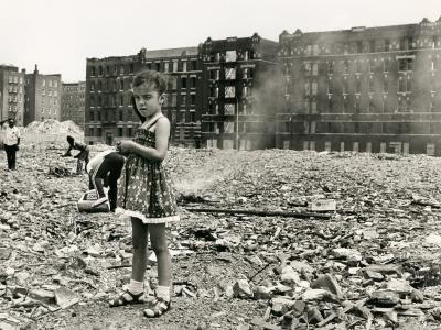 Black and white photograph. The photograph captures a place where a building use to be, but now there is only dust and rumble. There is what looks be a large apartment building in the back ground and other buildings behind it to the left.  In the left foreground there is a young girl in a dress squinting at the camera. She's wearing socks and sandals. There are three men behind her gathering things from the building rubble. 