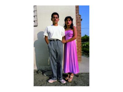 Color photograph. Image of a young man and a young woman standing at a street corner. The side of a building is behind them. To the right you can see slightly down the sidewalk. The young man is wearing a white t-shirt and grey dress pants with a black belt and white converse tennis shoes. The young woman is wearing a pink shiny silk dress with white open toe heels on.