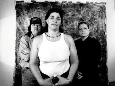 Black and white photograph. There are three feminine figures standing center, the view cuts off just below their waists. There is a backdrop taped up on the wall behind them. The figure in the center stands in from on the two people behind her, her hands hands rest on her belt buckle. She's wearing a white, tank top and black pants. The figure to the left has her arms crossed. She's wearing a long sleeve shirt and a black baseball hat that reads "DUKE". The figure to the right is wearing all black.