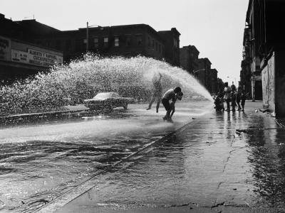 Black and white photograph of a city street with buildings framing the street on either side. In the middle ground there is a spraying arch of water from a fire hydrant. Two younger figures are jumping up into the arch of water. 