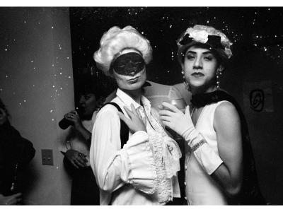 Black and white photograph. The image has two central figures hugging from the side and posing for the camera. Both figures are dressed up. The left figure is wearing a black, decorative face mask and a long sleeve white ruffle-y shirt. The right figure is wearing a decorative hat with a bow, tulle and flowers. They are also wearing white elbow length gloves, large dangly peal earrings, a glittery bracelet over the right glove that holds a drink, and  a white vest.