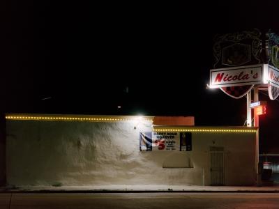 Color photograph. Image of a side of a building painted white with yellow lights lining the top edge, illuminated at night. Other neon signs say "cocktails" and "open." A large sign reads  "Nicola's" at the top of a metal street sign pole.