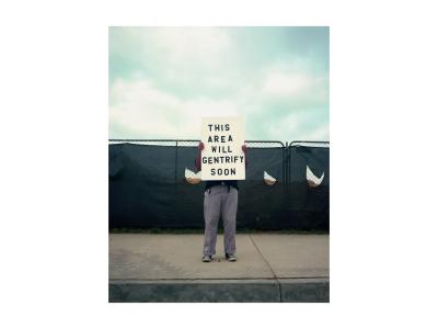 Color photograph. In the middle ground there is a person standing on a sidewalk holding a medium-sized sign in front of their face and torso. The sign reads: "This area will gentrify soon". The person is wearing grey pants rolled up at the cuffs, a black t-shirt, a black belt, black converse tennis shoes and there is a set of keys hanging by the left belt loop. There is a cloudy sky above the figure and a metal construction fence with black construction blind fabric blocking the view to the building site.