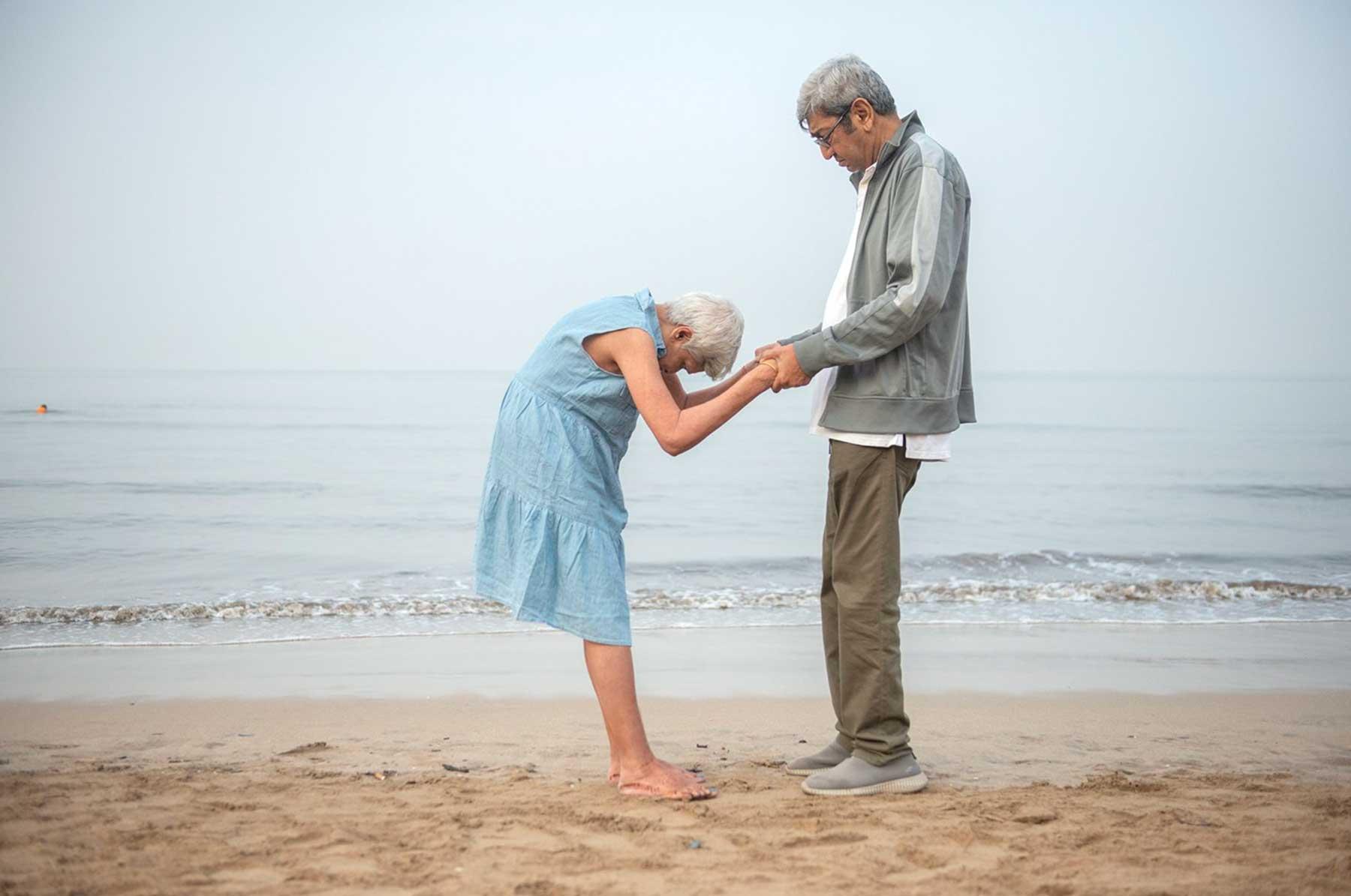 Photo of woman and man standing on a beach facing eachother, the women in bowing her head and the couple is clasping one another's hands r© Manjari Sharma, 2020.