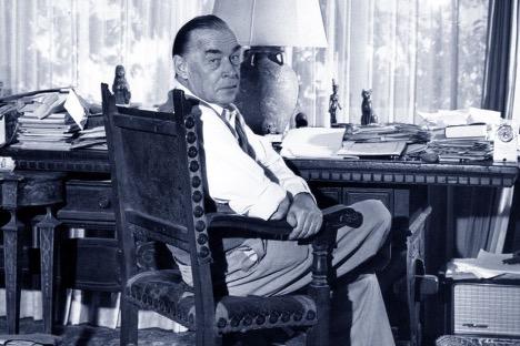 Black and white photo of Erich Maria Remarque sitting in a chair at a desk. His head is turned over his right shoulder to look at the camera